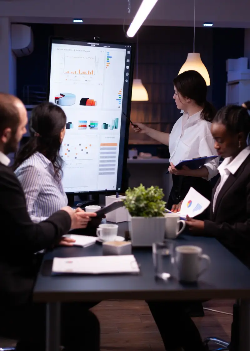 A group of professionals engages in a meeting, analyzing charts and data displayed on a digital screen. Coffee and papers are on the table.