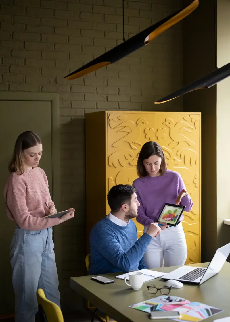 A group collaborates in a cozy workspace, discussing a colorful design on a tablet, with a laptop and notepad on the table.